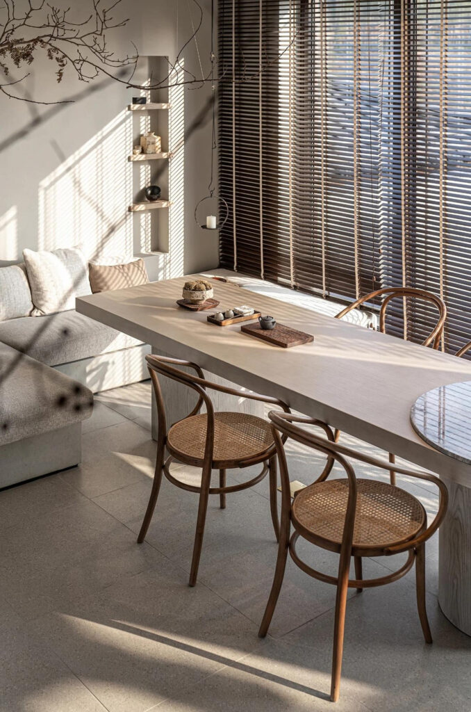 Moderni ruokailutila, jossa vaalea pöytä ja puisia tuoleja. Auringonvalo siivilöityy sisään ikkunan sälekaihtimien läpi.